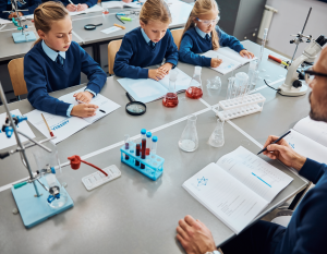 Three primary-aged children in school uniform sit at a science classroom table with beakers, test tubes and a microscope, writing in their notebooks while a teacher supports them.