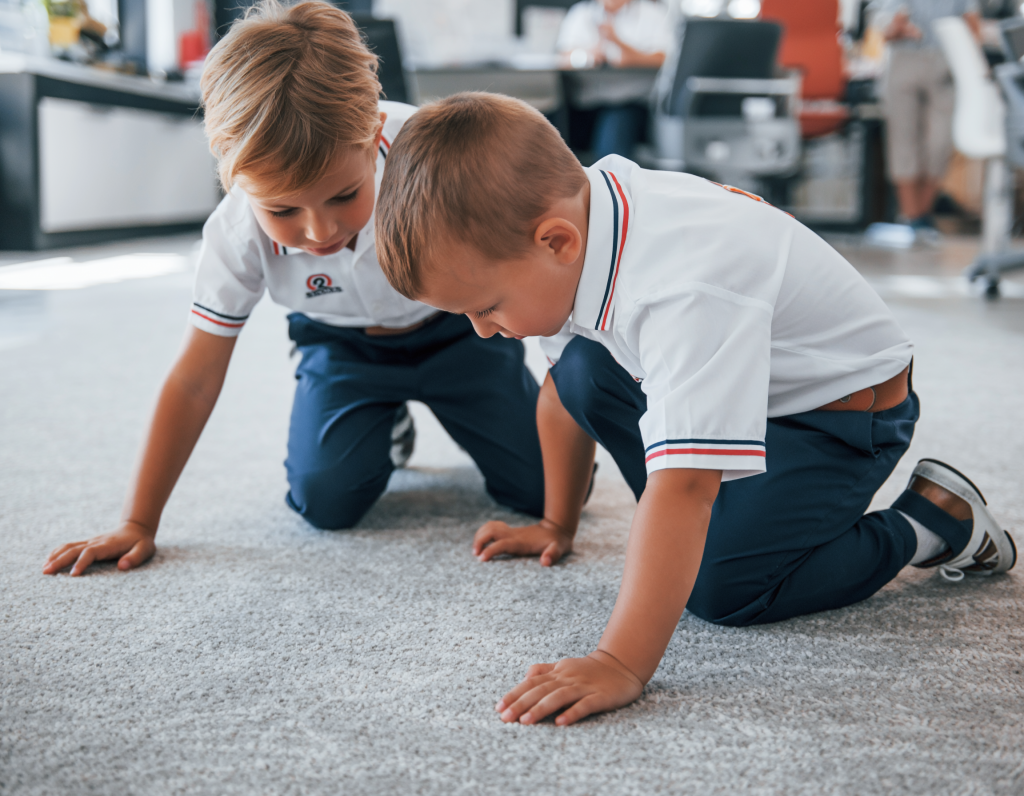 Two young children in school uniform kneel on a classroom carpet, focused on an activity on the floor, with a blurred classroom in the background.