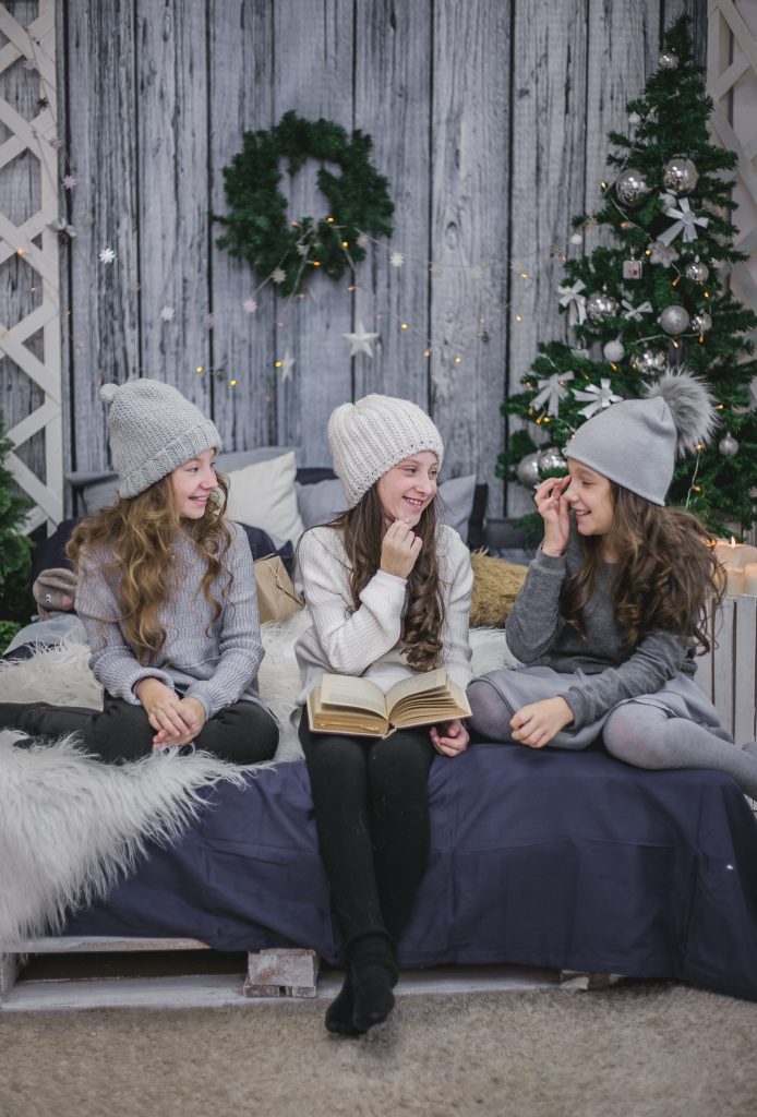 Three children wearing knitted winter hats sit on a bed indoors, smiling and talking while one holds an open book; a decorated Christmas tree and wreath are in the background.