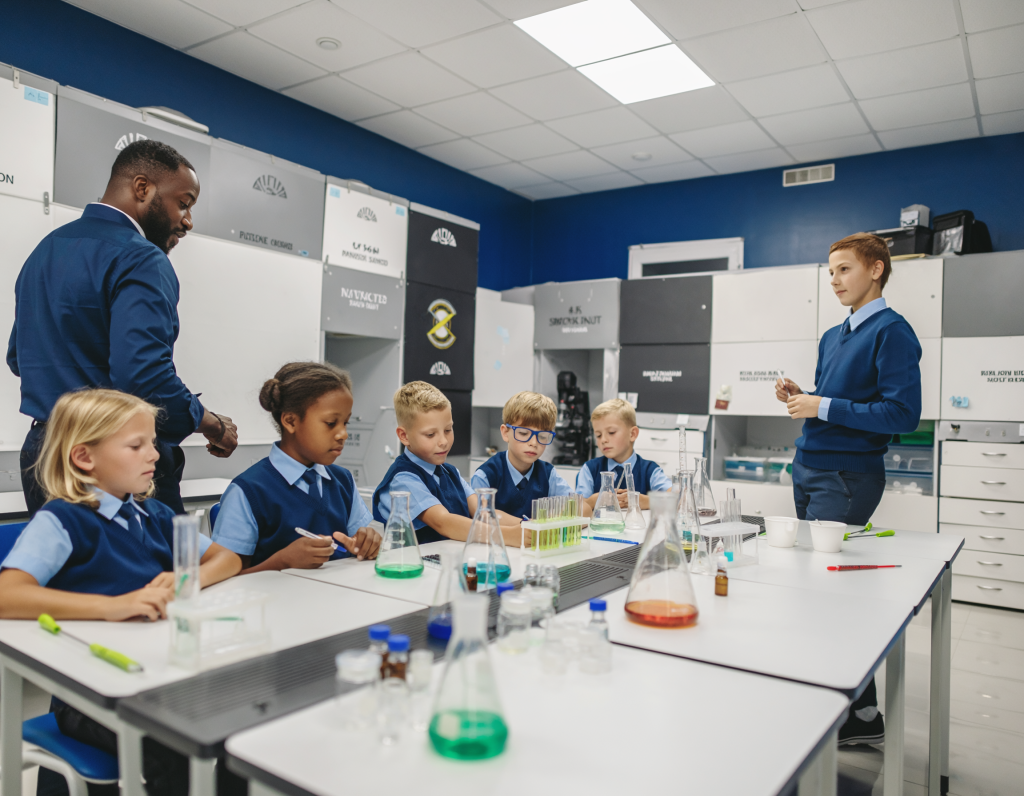 A group of primary school children in blue uniforms sit around a table in a science classroom, working with beakers and flasks of coloured liquids while a teacher supervises.